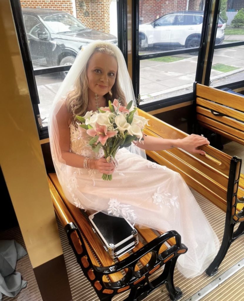 A bride in a white wedding dress and veil sits on a wooden bench inside a PST Trolley, holding a bouquet of flowers